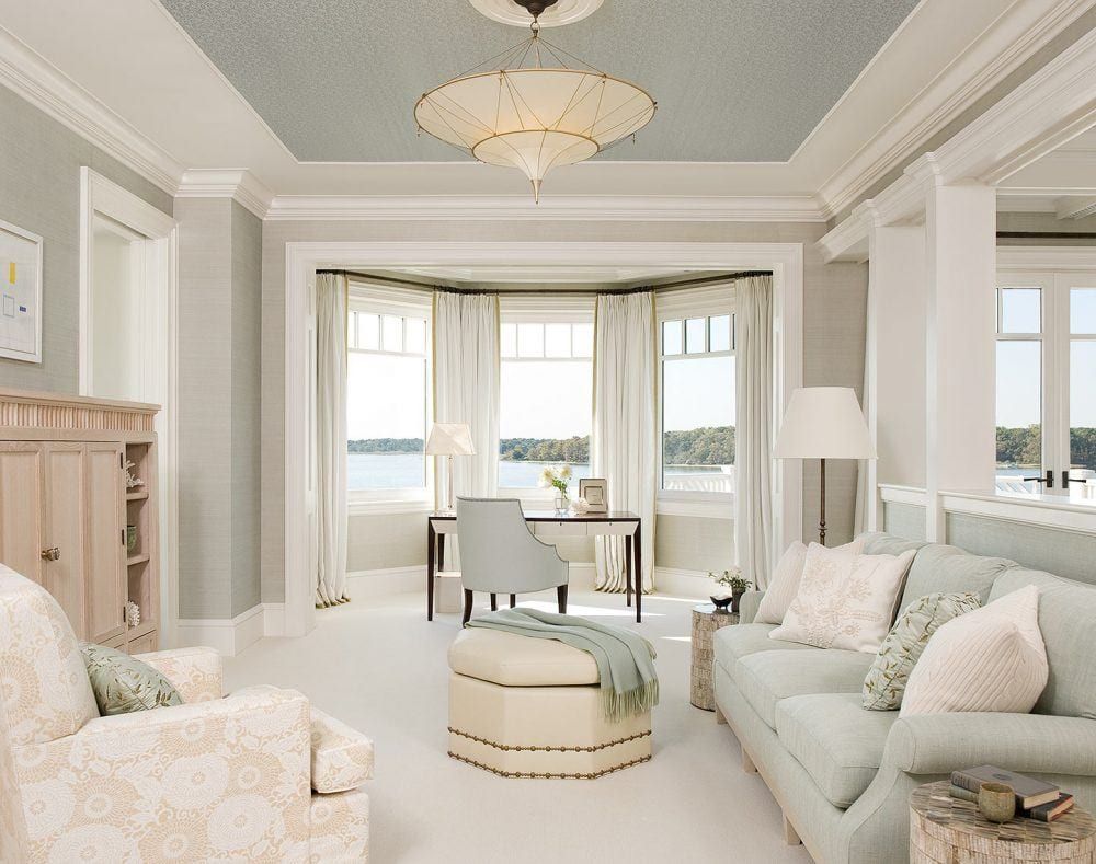 Elegant bay window study with cream curtains framing a lake view, light blue sofa, and classic chandelier.