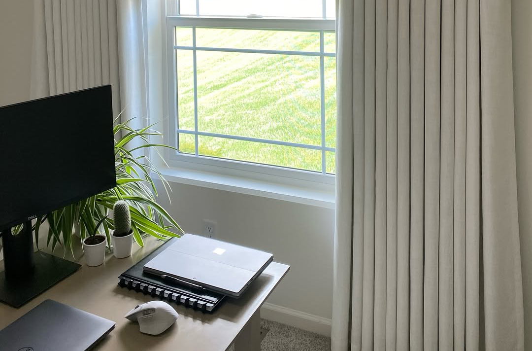 Cozy home office corner featuring a computer desk, houseplants, and modern white pleated curtains.